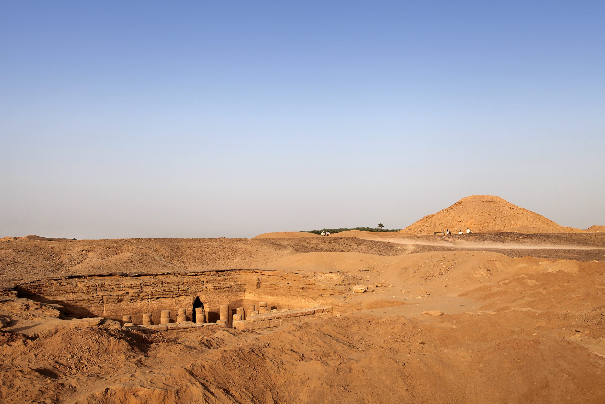 El-Kurru archaeological site, featuring a recessed temple and a short, blunt pyramid, under a clear, blue sky.